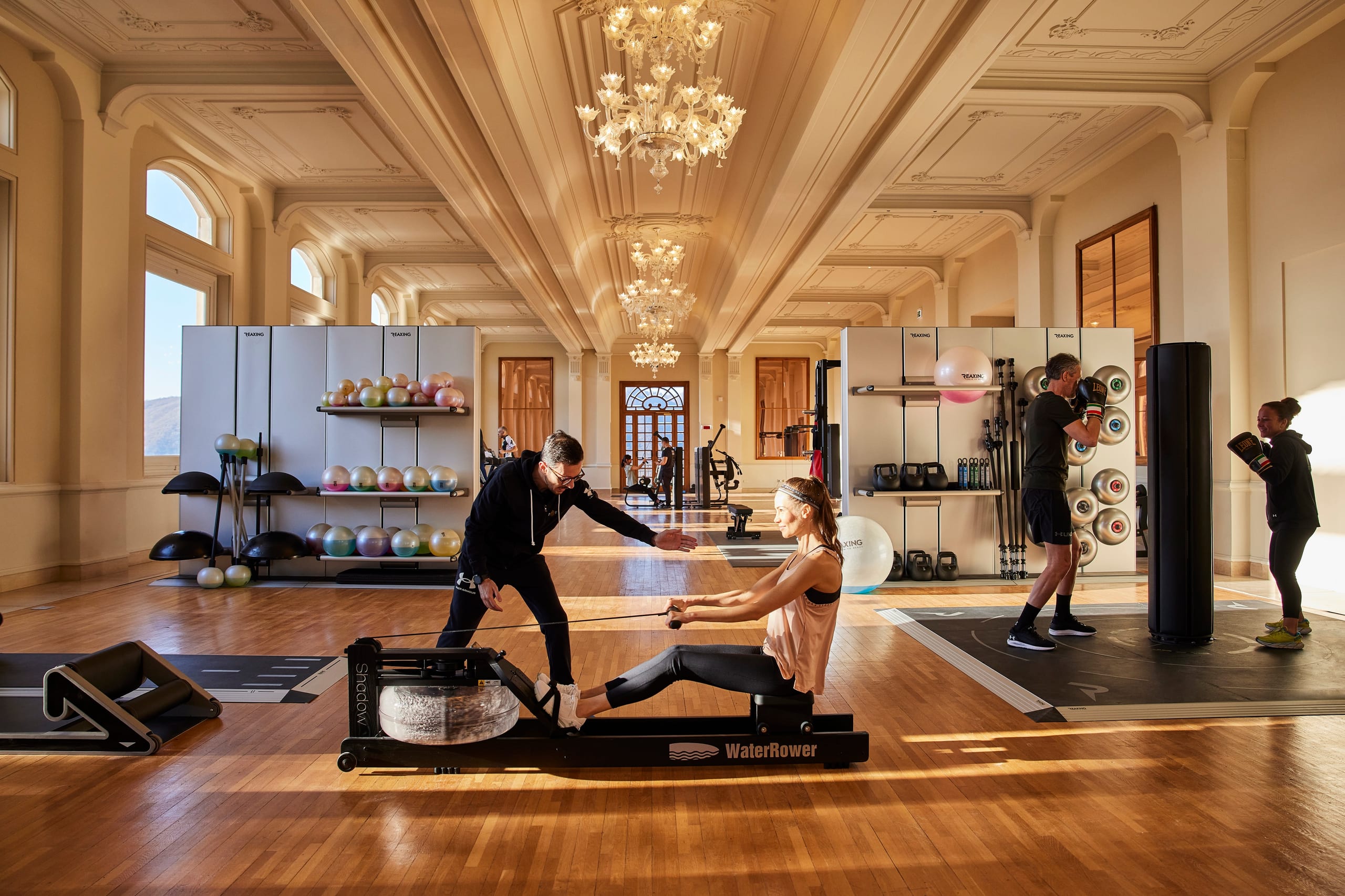Female guest enjoying a fitness coaching session using a water rowing machine, and male guest practicing fitness boxing, in the Ballroom of Palazzo Fiuggi turned into a gym