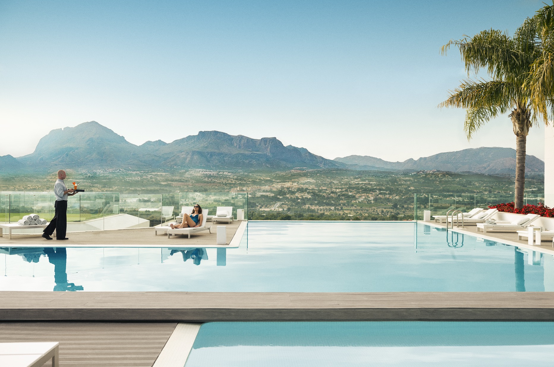 Female guest relaxing by the pool at SHA Wellness Clinic while a waiter brings her a healthy juice, with mountains in the background
